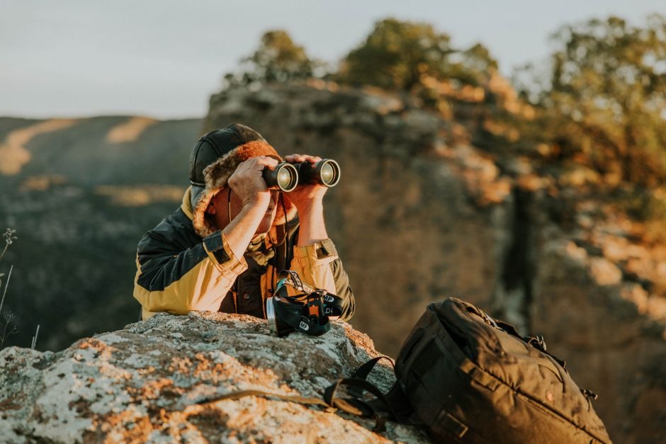 Outdoor observer using binoculars with a camera hanging at their side on a rocky overlook.